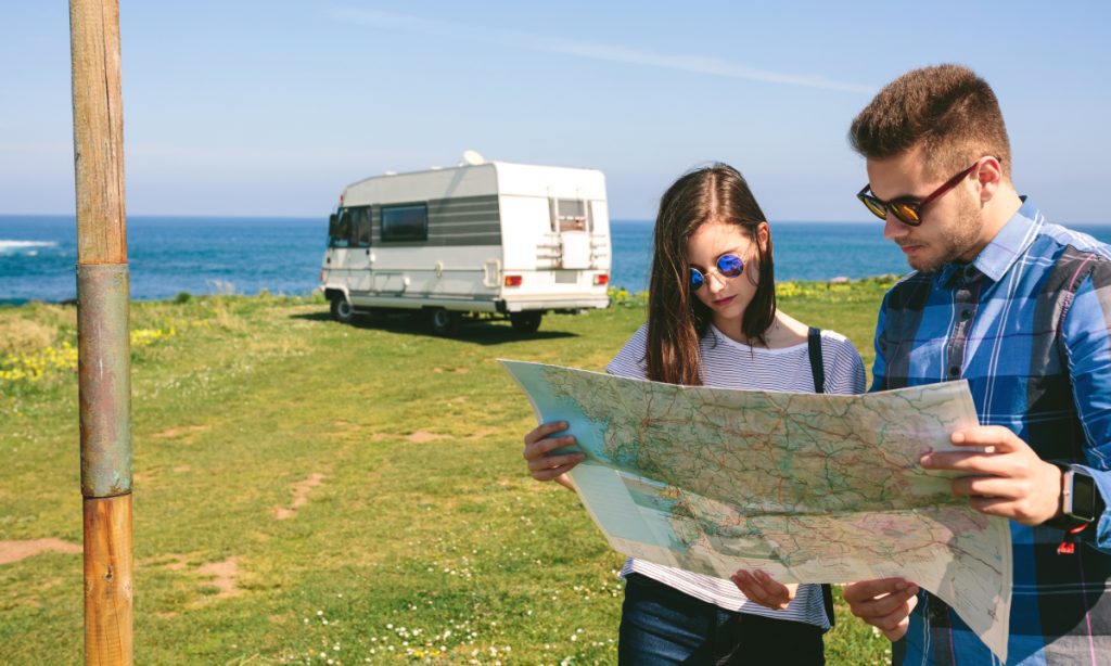 Una joven pareja consulta un mapa cerca de la costa con una autocaravana al fondo.
